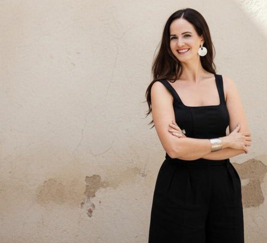 Smiling woman in black outfit standing against a beige wall.