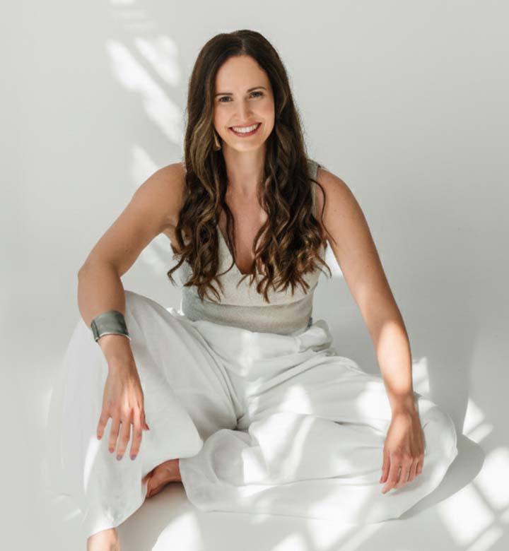 A woman with long dark hair smiles while sitting on the floor in white clothing.